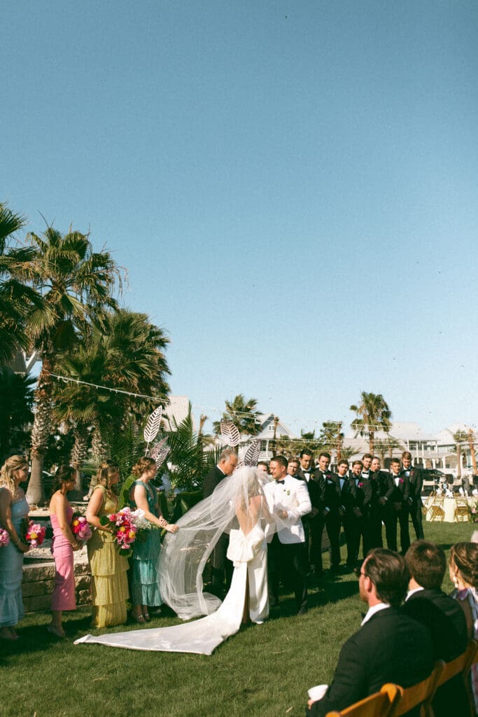 View of an outdoor wedding ceremony with a bright blue sky and palm trees in the background. The bride and groom are holding hands at the alter and the bridesmaids are wearing bright colorful dresses that fit the tropical setting. Photo taken by Lillian Jean Photography a Washington based wedding and elopement photographer who specializes in lifestyle wedding photography. 