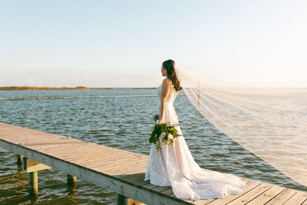 Bride stands on a wooden pier overlooking a calm bay. Her veil is floating on the wind and drifts out of frame. Photo taken by Lillian Jean Photography a Washington based wedding and elopement photographer who specializes in lifestyle wedding photography. 