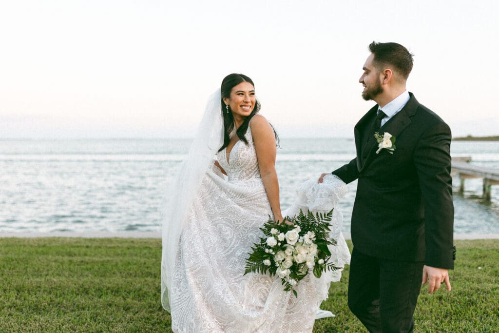 Candid photo of bride and groom walking together. Calm bay can be seen in the background. The bride is slightly in front of the groom looking back at him with a large smile. The groom is holding the train of her dress. Photo taken by Lillian Jean Photography a Washington based wedding and elopement photographer who specializes in lifestyle wedding photography. 
