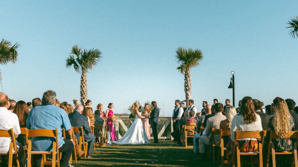 Wedding ceremony scene in a Tropical setting with palm trees and bright blue skies. Bride and groom are at the alter holding hands. Photo taken by Lillian Jean Photography a wedding and elopement photographer based out of Washington State and available worldwide.